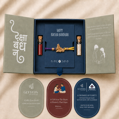 A Raksha Bandhan gift set displayed in an open sage green box on a beige fabric background. The set includes a navy blue card reading 'HAPPY RAKSHA BANDHAN' with a gold-toned cord ornament, two small vials, and three oval cards below.